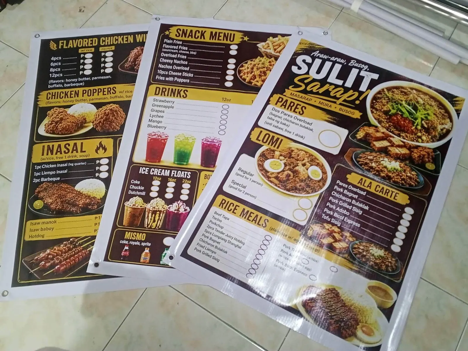 Three food-stall menu tarpaulins laid on a tile floor